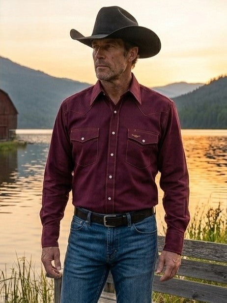 A man wearing a burgundy canvas western long-sleeve, American Made snap shirt, blue jeans, and a cowboy hat, standing outdoors in front of a barn with a lake in the background. Made by Ruddock Shirts in El Paso, Texas, Made in USA Flying R Ranchwear since 1955