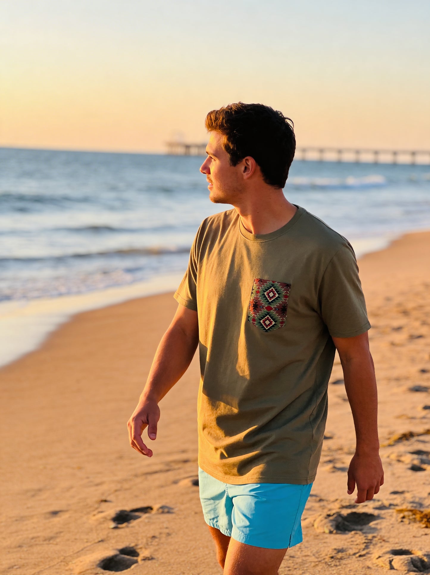 Man walking on a beach at sunset wearing a green t-shirt and blue shorts.