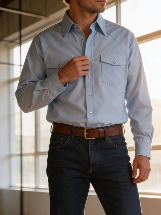 Man wearing a light blue button-up shirt and dark jeans indoors with large windows.