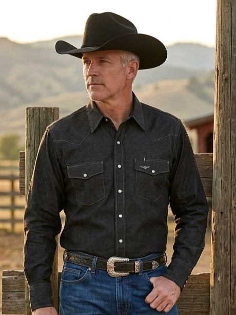A man wearing a black western long-sleeve, American Made snap shirt, blue jeans, and a black cowboy hat, standing outdoors in front of a wooden fence with hills in the background. Made by Ruddock Shirts in El Paso, Texas, Made in USA Flying R Ranchwear since 1955