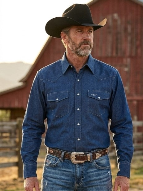 A man wearing a blue denim western long-sleeve, American Made snap shirt, blue jeans, and a black cowboy hat, standing outdoors in front of a wooden fence with hills in the background. Made by Ruddock Shirts in El Paso, Texas, Made in USA Flying R Ranchwear since 1955