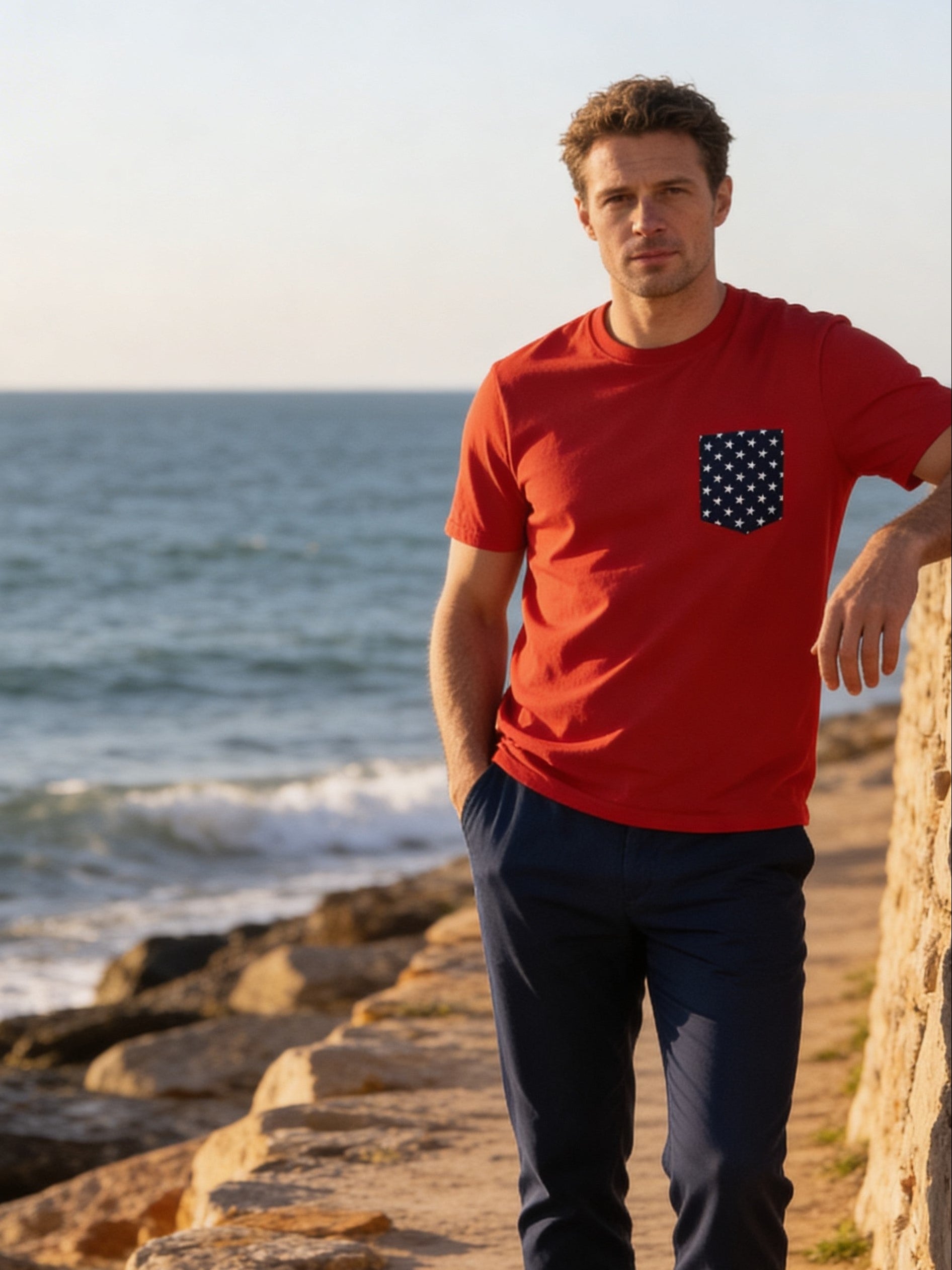 Man in a red shirt standing on a rocky beach with ocean and sailboat in the background